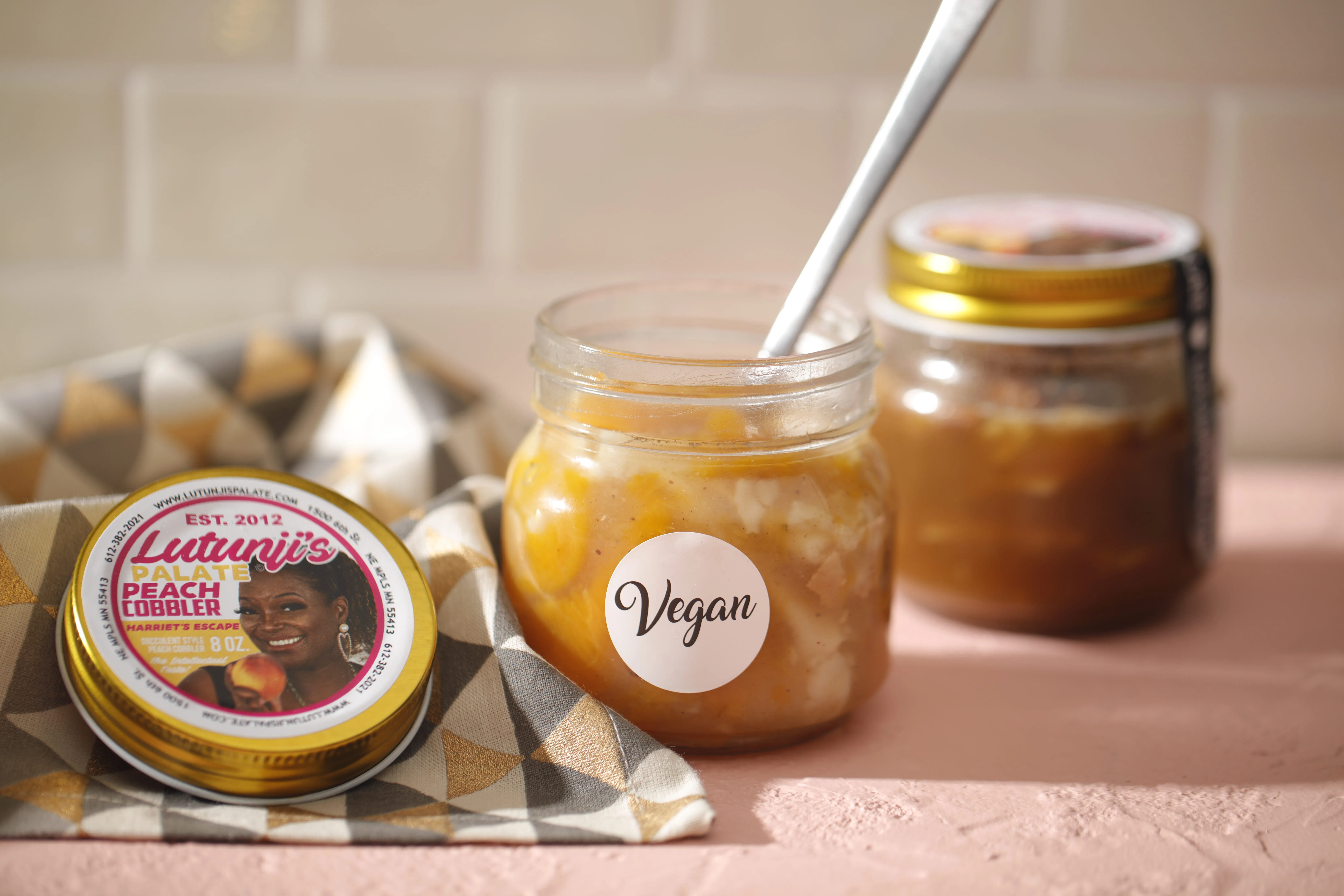 Lutunji's Palate Peach Cobbler in a glass jar with a spoon in it and the lid from the jar to the left of the jar with the Lutunji's Palate logo on it. The lid is sitting on a patterned linen. There is also a jar of Lutunji's Palate Pecan Cobbler in the back right of the photo. It is slightly out of focus.